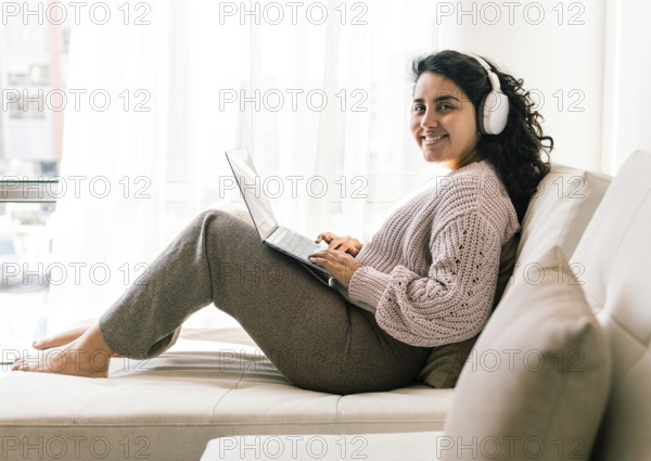 A young latin woman in cozy attire sits comfortably on a sofa at home, using a laptop with