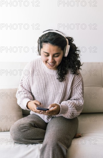 A young latin woman sits comfortably on a couch, wearing headphones and looking at her phone She