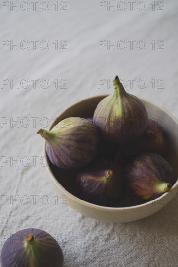 A rustic ceramic bowl filled with ripe, purple figs placed on a textured linen tablecloth,