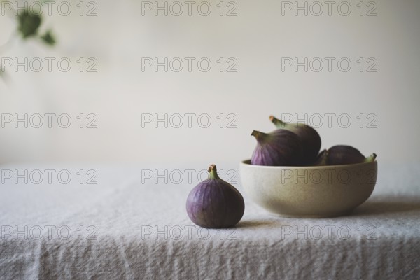 A collection of ripe figs in a ceramic bowl on a rustic tablecloth One fig is placed outside the