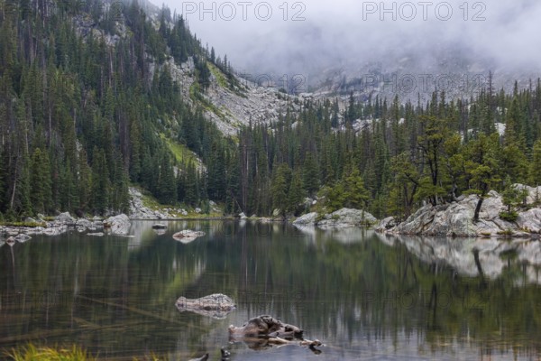 Misty atmosphere blankets a peaceful lake surrounded by dense forests and rugged mountains in the