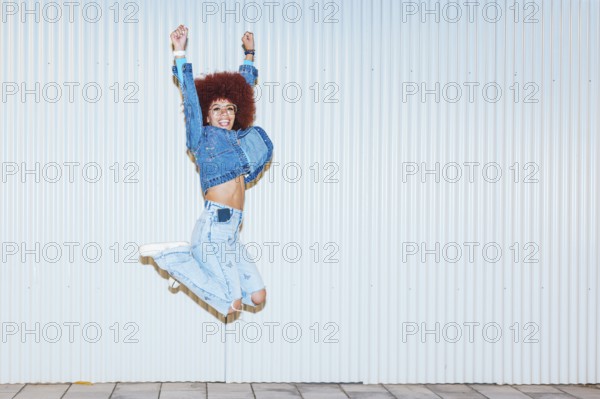 A joyful African descent woman with afro hair and fashionable denim outfit jumping energetically