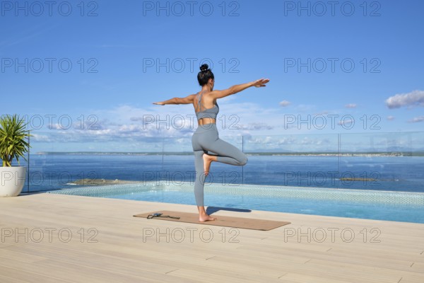 A woman practices yoga on a deck overlooking the ocean. She is in a balance pose, embracing