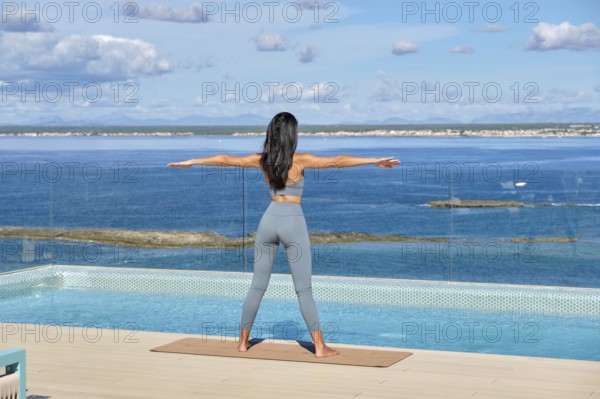 A woman stands on a yoga mat by an infinity pool, facing the ocean, arms extended in a peaceful