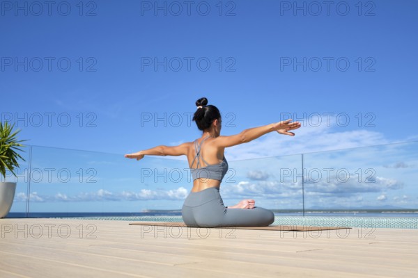 A woman in a gray yoga outfit practices yoga on a deck overlooking the ocean. Her arms are