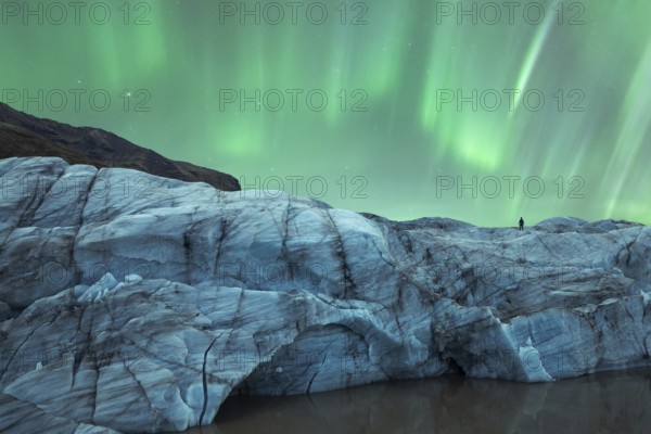 A solitary figure stands atop a glacier in Iceland, gazing at the mesmerizing Northern Lights that