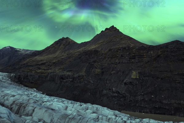 The Northern Lights cast a radiant glow over rugged mountains and a glacial landscape in Iceland,