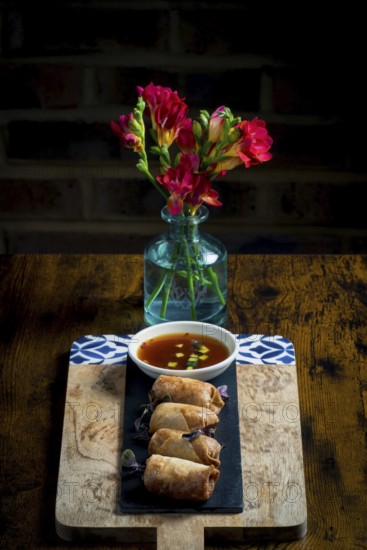 A wooden board holds crispy spring rolls beside a bowl of dipping sauce, set against a vibrant