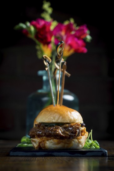 A gourmet pulled pork slider presented elegantly with colorful flowers in the background The food