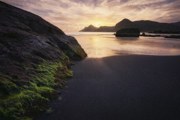 The tranquil sunset casts a golden hue over the rugged rocks and calm waters of Cala del Lastre in