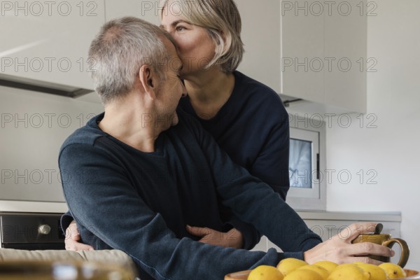 A senior couple enjoys a tender moment in their kitchen One person kisses the other on the head,
