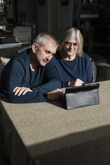 A senior couple sits at a table in a cozy living room, engaged with a digital tablet They appear to