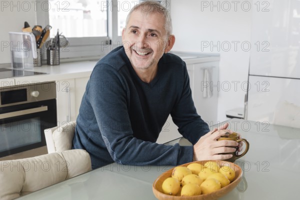 A cheerful senior man sits in a bright kitchen, holding a mug, with a basket of lemons nearby The