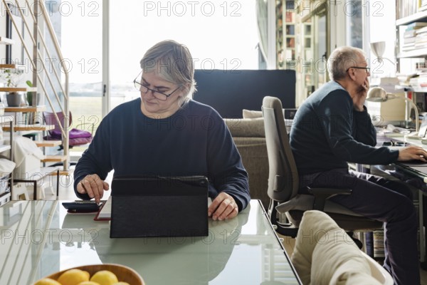 A senior couple sits in a bright home office, one using a tablet, the other focusing on a computer