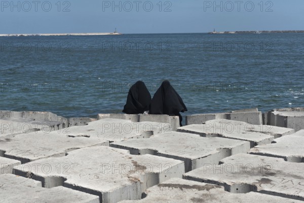 Two Muslim women wearing black abaya or chador sit on the concrete breakwater on the Mediterranean