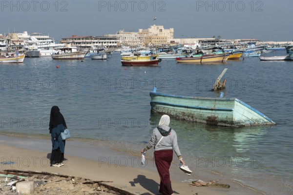 Alexandria, Egypt. December 1st 2022 Two Muslim women walk on the beach near a sunken boat in front