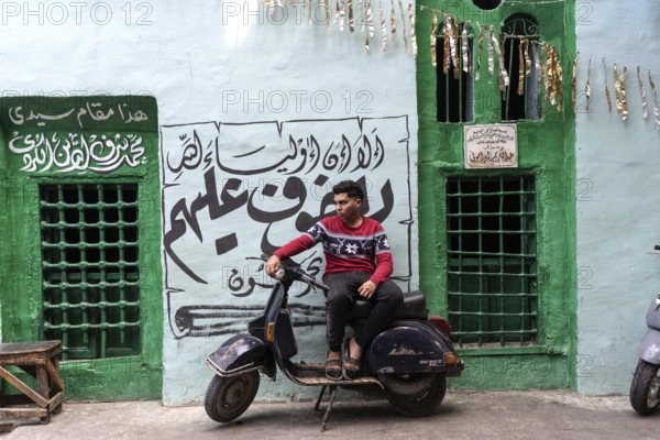 Cairo, Egypt. April 15th 2025. A young Egyptian man sits on a motor scooter outside a brightly