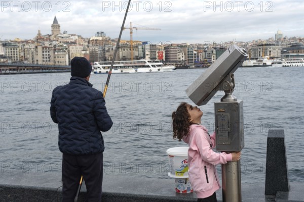 Istanbul, Turkey. January 3rd 2025. A man fishing and a child plays with street binoculars beside