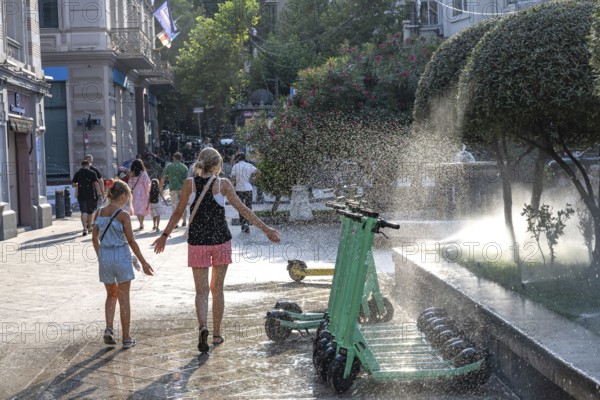 Tbilisi, Georgia. July 29th 2025. A woman and child enjoy the cool spray of a garden sprinkler in