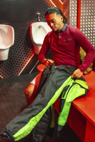 Young man sitting in a changing room, putting on a black and green racing suit for an upcoming