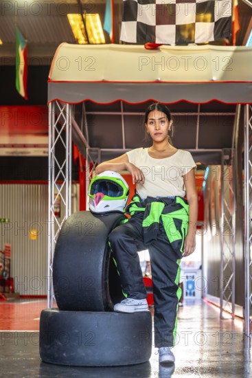 Woman wearing a racing suit and helmet standing confidently by tires and checkered flags at an