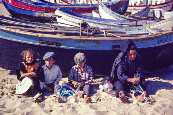 Fisherman and children repairing nets on beach by traditional fishing boats, Nazaré, Portugal,