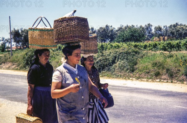 Three women walking on street in rural area carrying wicker baskets on their head, Portugal, Europe
