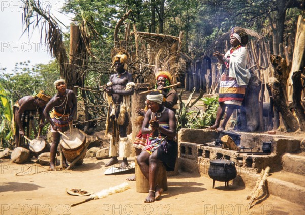 Group of Zulu people dressed in traditional attire, performing an outdoor ceremony at a village,