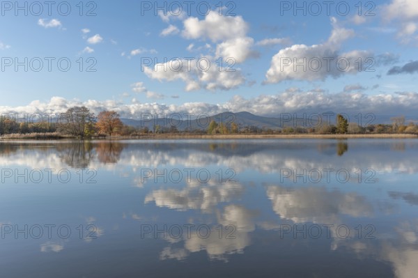 The river flows flat and reflects clouds and mountains under a clear sky. Autumn trees draw the
