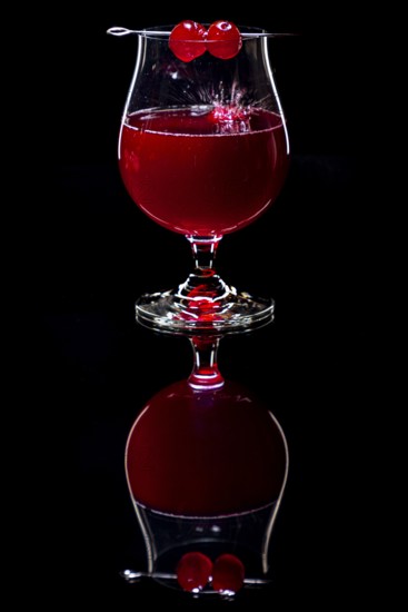Cherry falls splashing into a cognac glass with a red drink against a dark background and