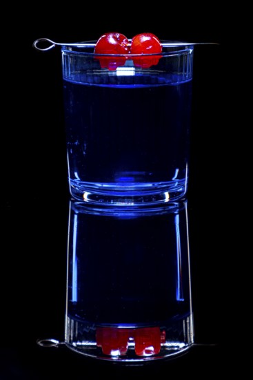 Blue drink in a glass with cherries, against black background and reflection