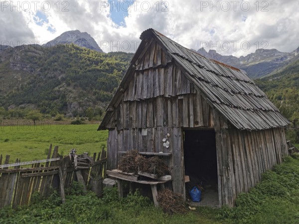 Rustic wooden cabin in a green meadow against an alpine mountain backdrop with cloudy sky, Peaks of