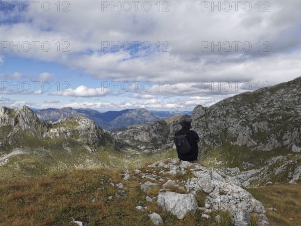 Person with backpack sitting on a rock and looking at a mountain panorama under cloudy sky, Peaks