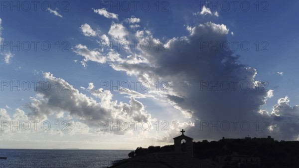 Dramatic sky with clouds and sunlight over sea with church silhouette in foreground, Himare,