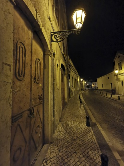Night view of a lonely paved street with cozy glowing lanterns and historic buildings, Faro,
