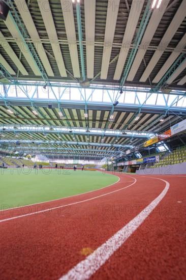 Interior view of a modern sports hall with red running track and green area under steel girders,