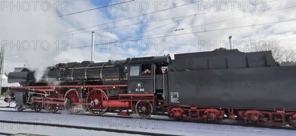 A black, historic steam locomotive emits smoke and stands on snow-covered tracks in winter,