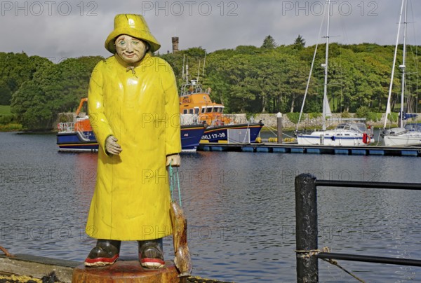 Statue of a fisherman in yellow raincoat at harbour in front of anchored boats with green hills in