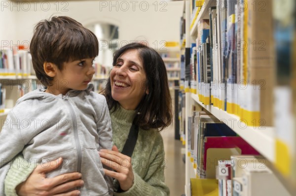 Mother holding her young child, both smiling and interacting happily while standing by shelves