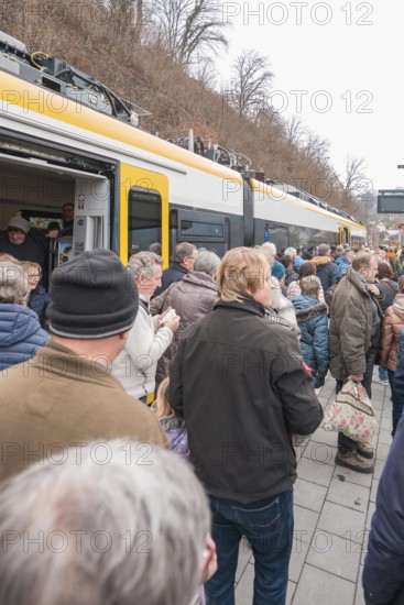 People board a yellow train standing at a platform in an urban area, opening of the Hermann Hesse