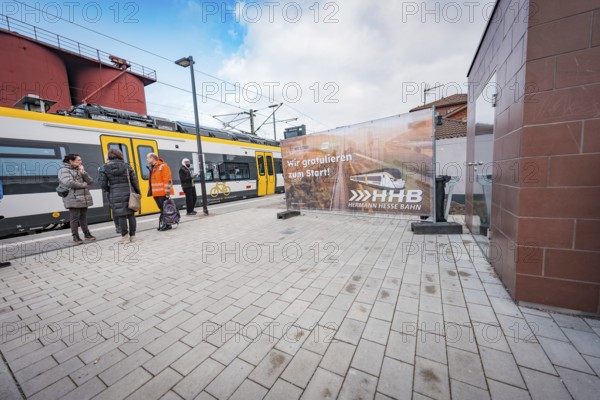 People wait on the railroad track next to a yellow train, a large poster is visible, battery