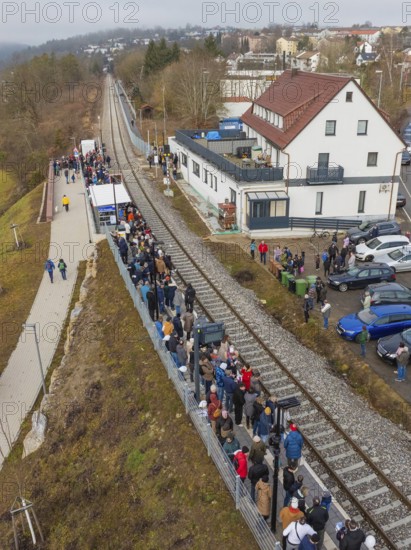 People wait for a train at a station along the tracks next to a road, opening of the Hermann Hesse