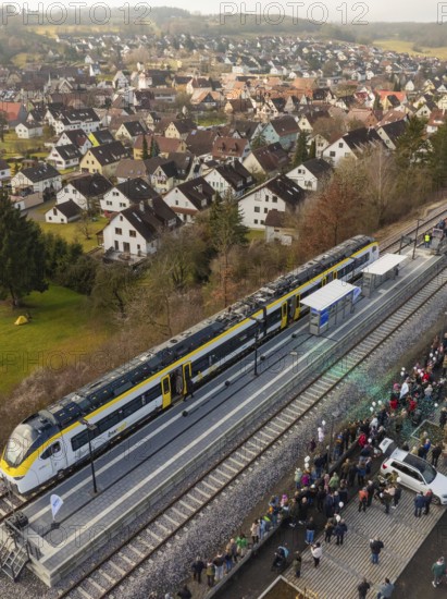 A train stops at the village station and many people are gathered. In the background, the village