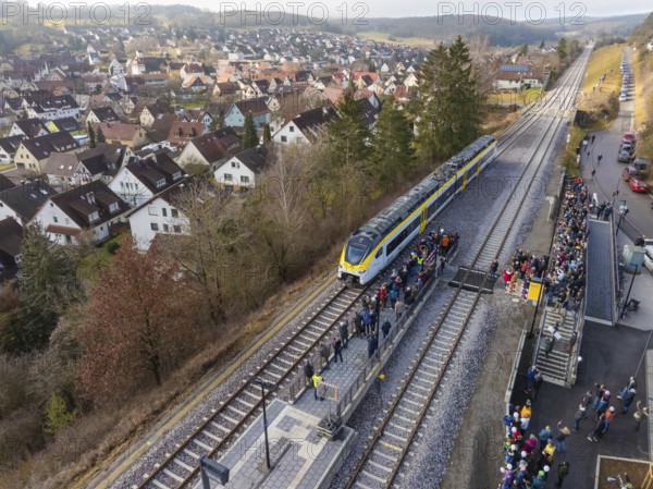 A train stops at a rural train station with surrounding people. The surrounding area shows a