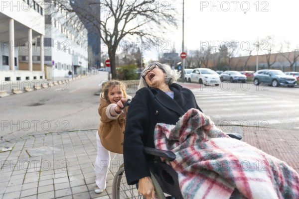 Granddaughter pushing her joyful grandmother in a wheelchair during an outdoor walk on a sunny day,