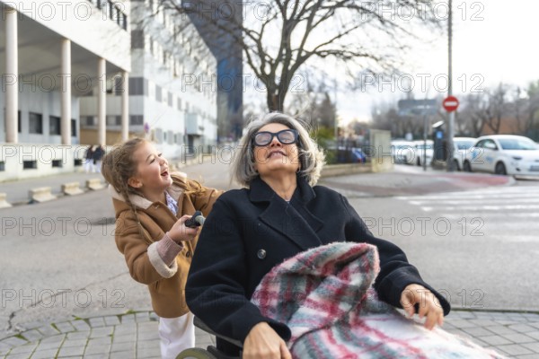 Happy granddaughter pushing senior grandmother in a wheelchair, enjoying a sunny day outdoors and