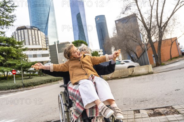 Happy granddaughter sitting on grandparent's lap in a wheelchair, holding hands and laughing with