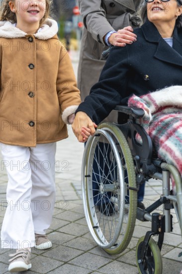 Granddaughter walking alongside her grandmother in a wheelchair, holding hands, with a woman