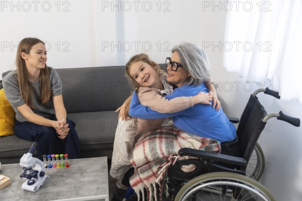 Small girl hugging her grandmother sitting in a wheelchair while a woman watches from the couch,