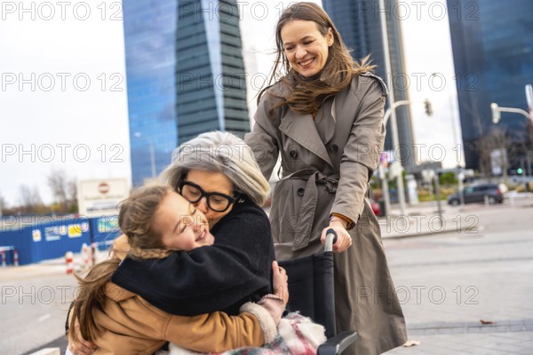 Granddaughter embraces her grandmother sitting in a wheelchair while a woman pushes them, enjoying